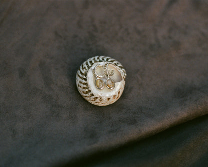 A shell with intricate design, holding two earrings on a textured fabric background