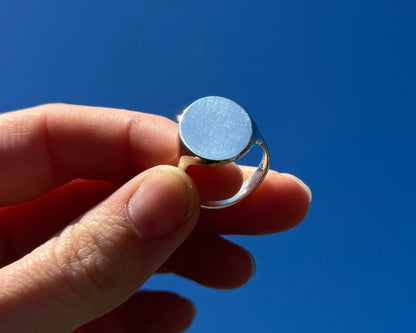 Hand holding a silver signet ring against a clear blue sky